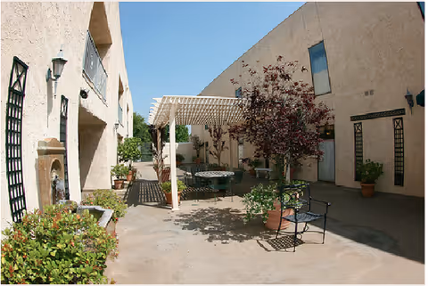Outdoor courtyard area at Evergreen Retirement Residence with a pergola providing shade over a round table and chairs. The courtyard is surrounded by beige stucco walls with windows and decorative elements. There are potted plants and small trees adding greenery to the space under a clear blue sky.