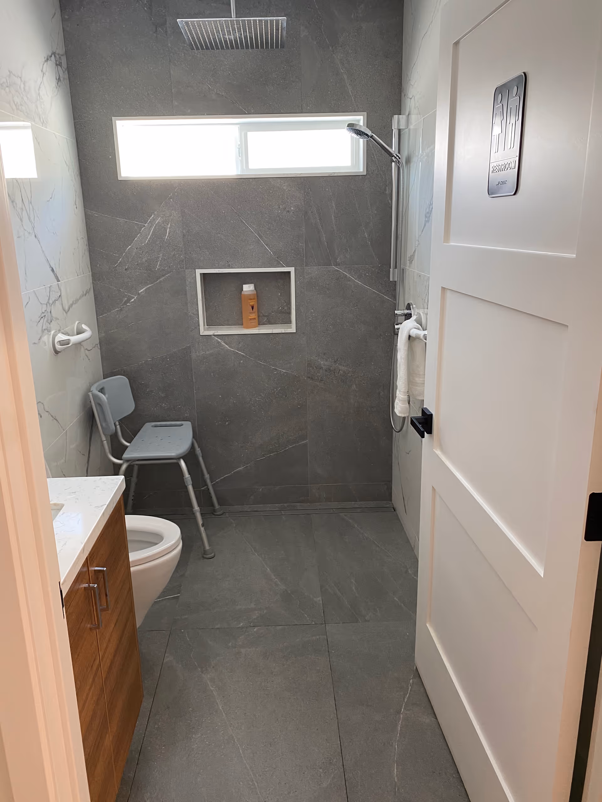 A modern bathroom with gray tiled floors and walls. There is a white toilet, a wooden cabinet with a white countertop, a gray shower chair, and a walk-in shower with a handheld showerhead. A small window is above the shower area, and a bottle of shampoo or soap is placed in a recessed shelf in the shower wall. The door has a sign indicating it is a restroom.