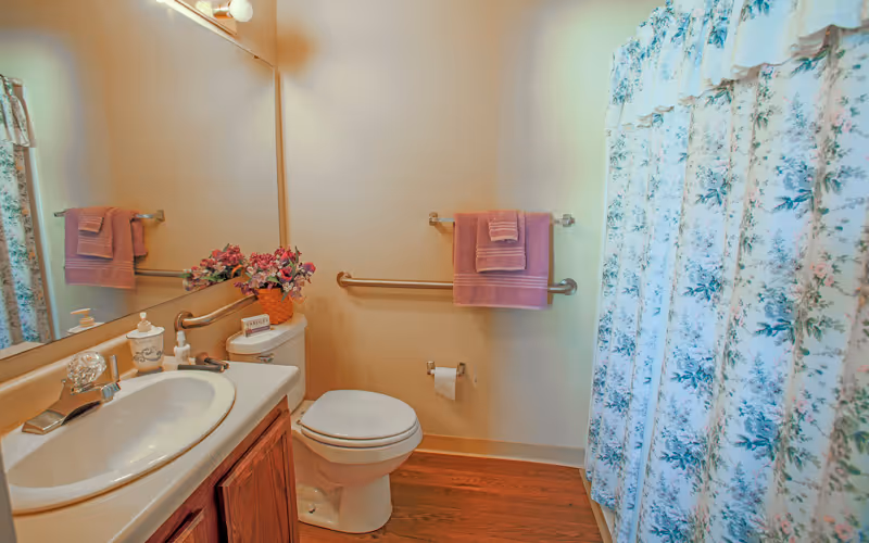 A bathroom with a white sink and countertop, a toilet, and a shower with a floral-patterned curtain. There are pink towels hanging on a grab bar above the toilet and on a towel rack. A large mirror is above the sink, reflecting part of the room. The floor is wooden, and there is a small flower arrangement on the toilet tank.