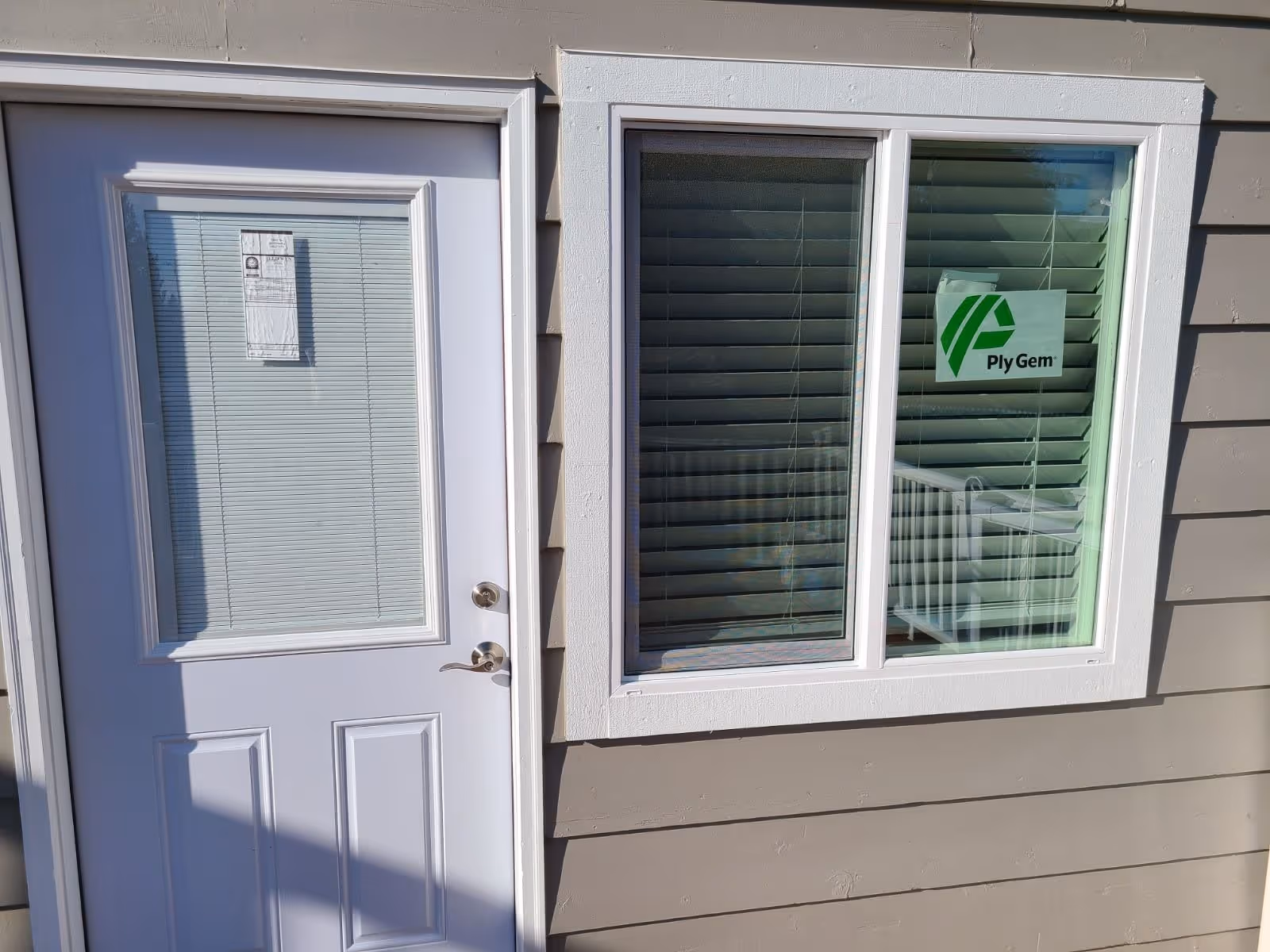 Exterior view of a beige building wall featuring a white door with a window covered by blinds and a window with white trim. The window has a green and white Ply Gem logo sticker on it.
