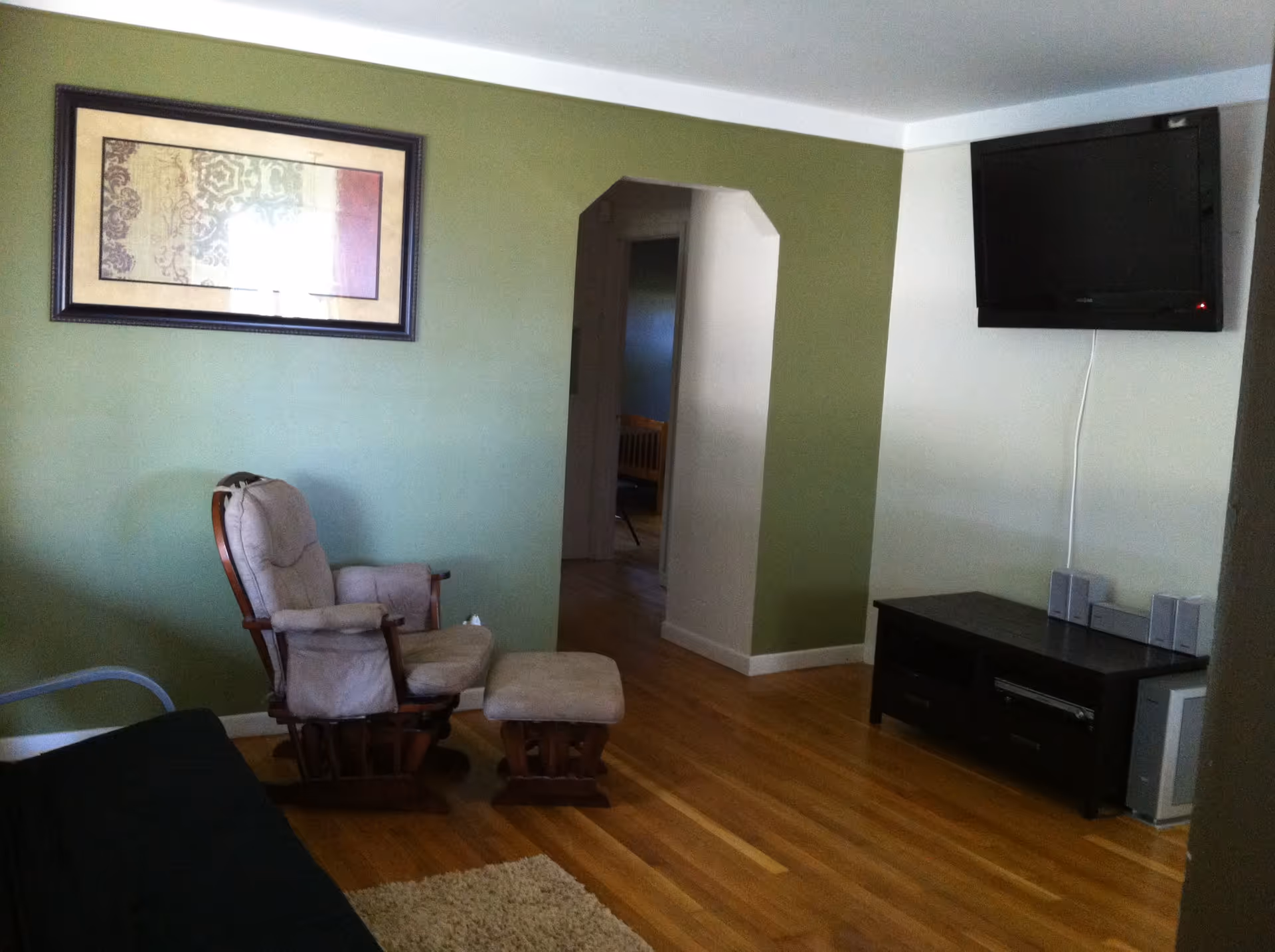 A living room with wooden flooring, a cushioned rocking chair with matching footrest, a black TV mounted on a white wall, a dark wooden TV stand with speakers and electronic devices, a green accent wall with a framed decorative artwork, and an open doorway leading to another room.