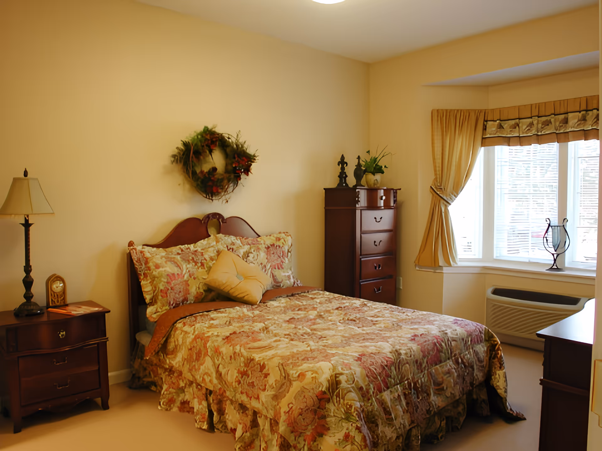 A cozy bedroom with a floral patterned bedspread on a wooden bed frame. There is a nightstand with a lamp and a clock on the left side of the bed. A decorative wreath hangs on the wall above the bed. To the right, there is a tall wooden dresser with decorative items on top and a window with beige curtains letting in natural light. An air conditioning unit is below the window.