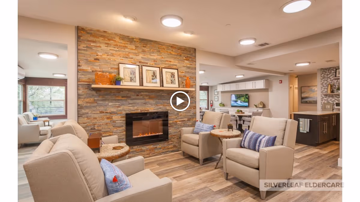 A cozy living room area in an eldercare facility featuring four beige armchairs with blue and white pillows arranged around a modern electric fireplace set in a stone accent wall. A wooden shelf above the fireplace holds framed artwork and decorative items. The room has wood flooring and recessed ceiling lights. In the background, there is a dining area with a TV mounted on the wall and a kitchen area with dark cabinetry and a towel hanging on the handle.