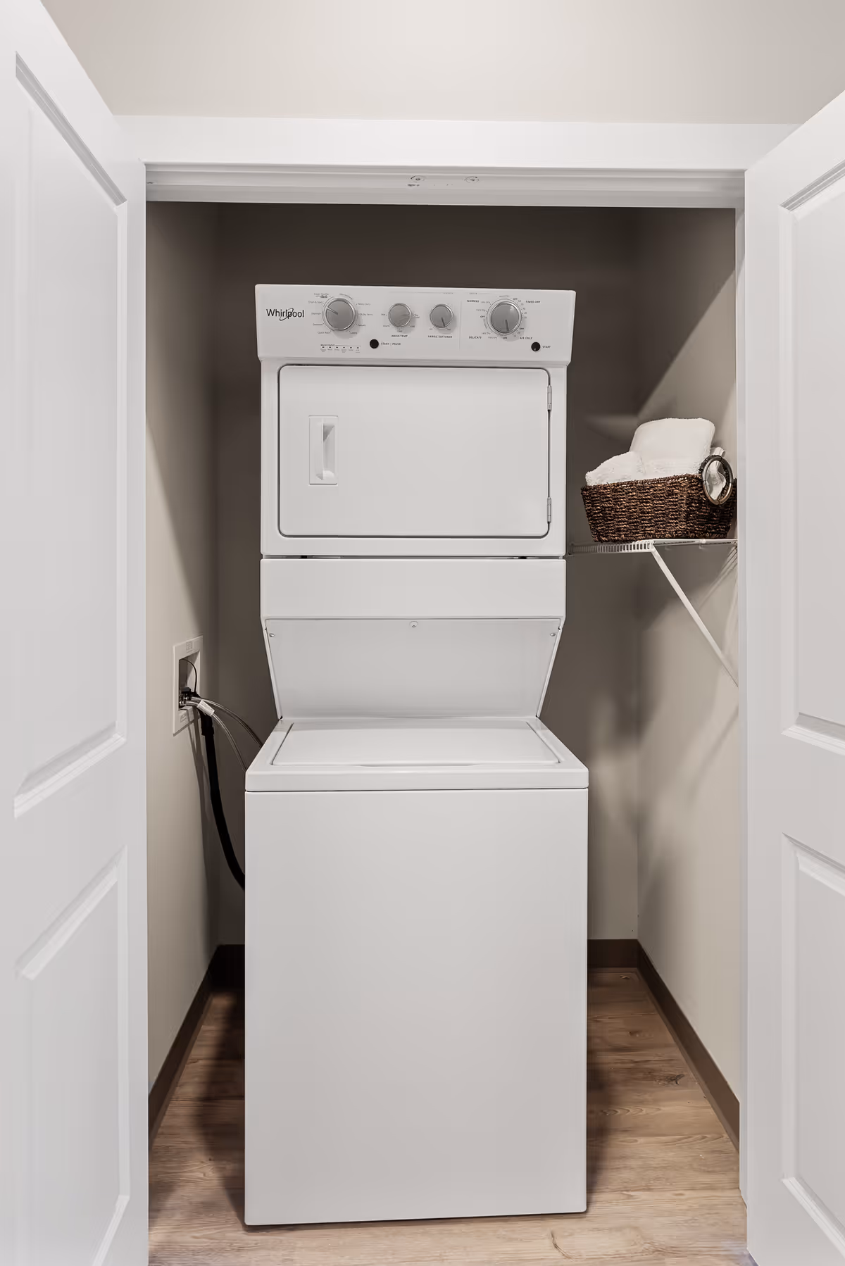 A compact laundry area with a stacked white Whirlpool washer and dryer unit inside a small closet with double doors. A wire shelf on the right holds a brown wicker basket with folded white towels and a small clock.
