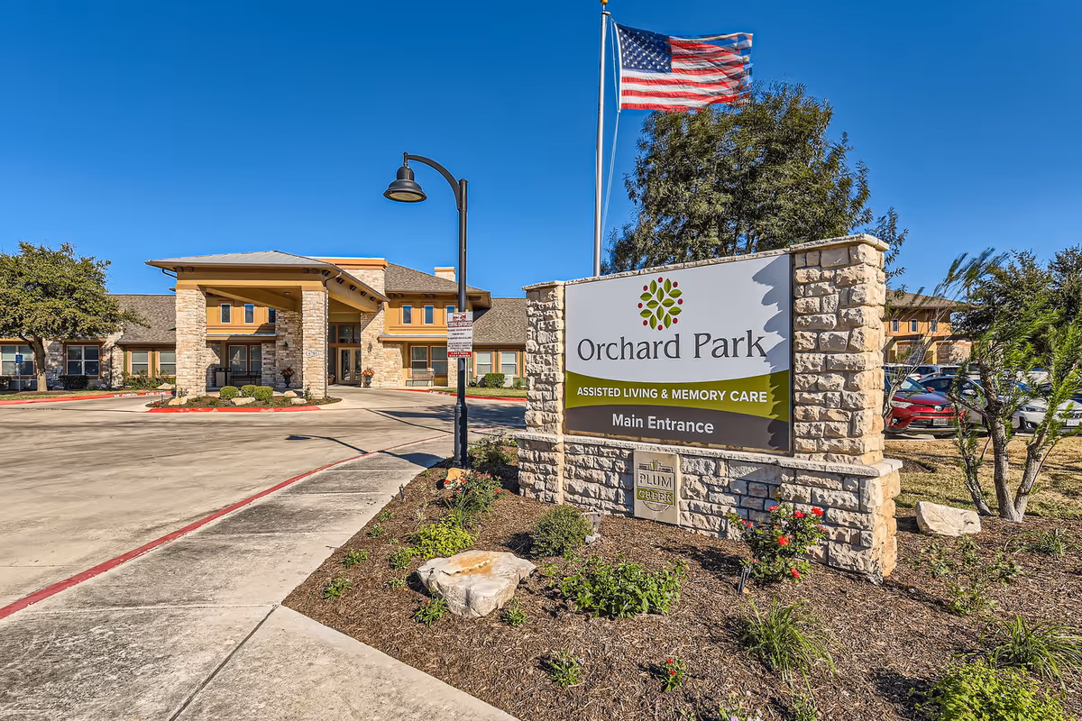 Exterior view of Orchard Park Assisted Living & Memory Care facility with a stone sign at the entrance, an American flag on a flagpole, landscaped plants, and a clear blue sky.