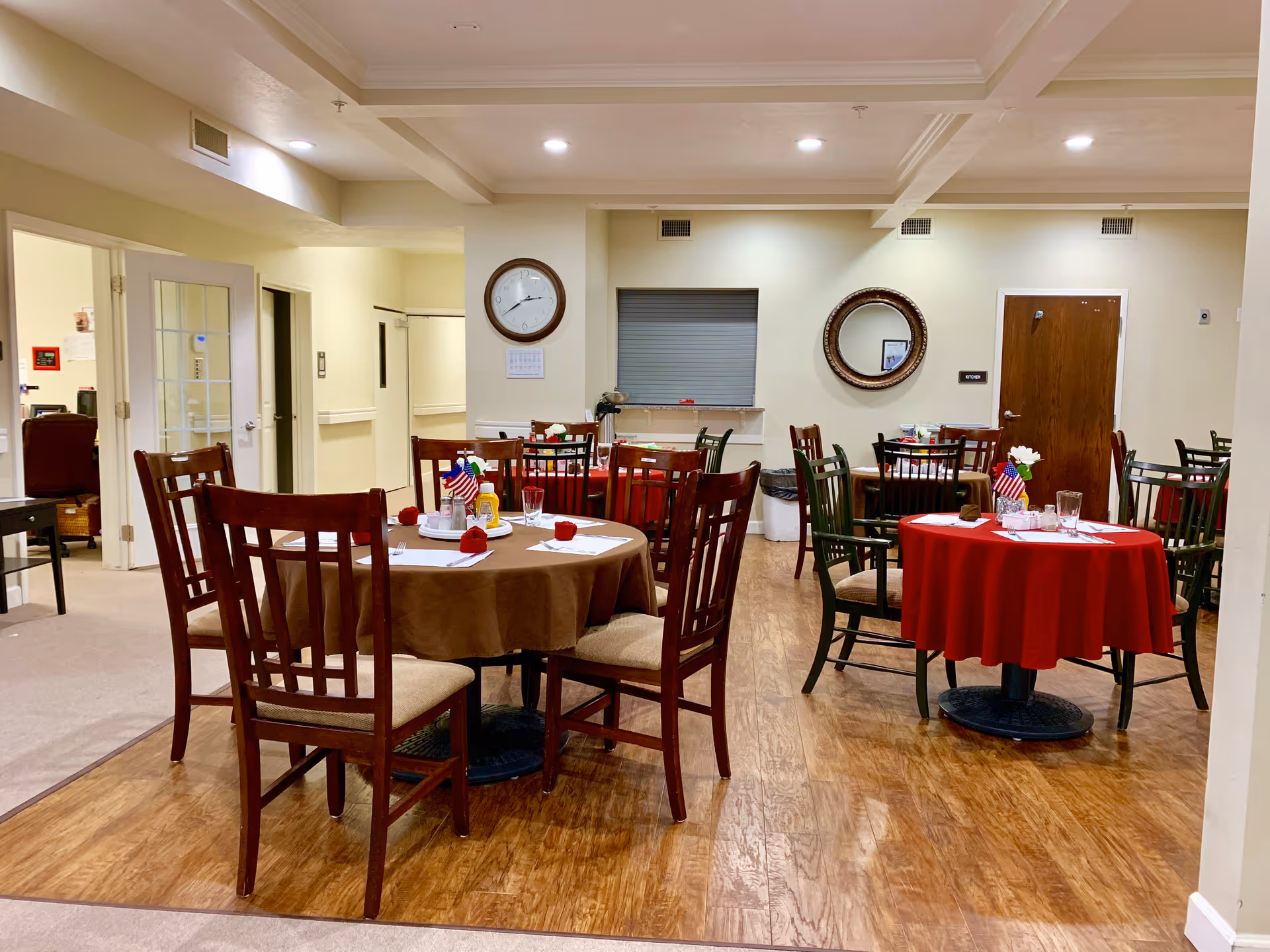 A dining area in an assisted living facility with several round tables covered with red and brown tablecloths. Each table is set with glasses, napkins, and small American flags. Wooden chairs with cushioned seats surround the tables. The room has wood flooring and cream-colored walls with a clock and a round mirror hanging on the wall. There are doors and an open doorway leading to other rooms.