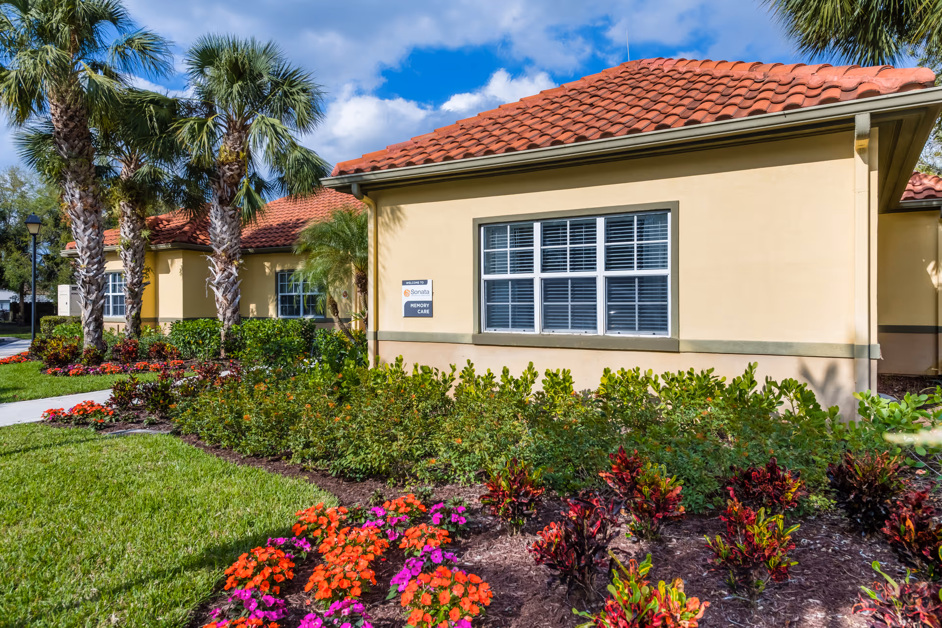 Exterior view of a single-story building with beige walls and a red tiled roof, surrounded by well-maintained landscaping including palm trees, green shrubs, and colorful flowers under a partly cloudy blue sky. A sign on the building reads 'Sonata Memory Care'.