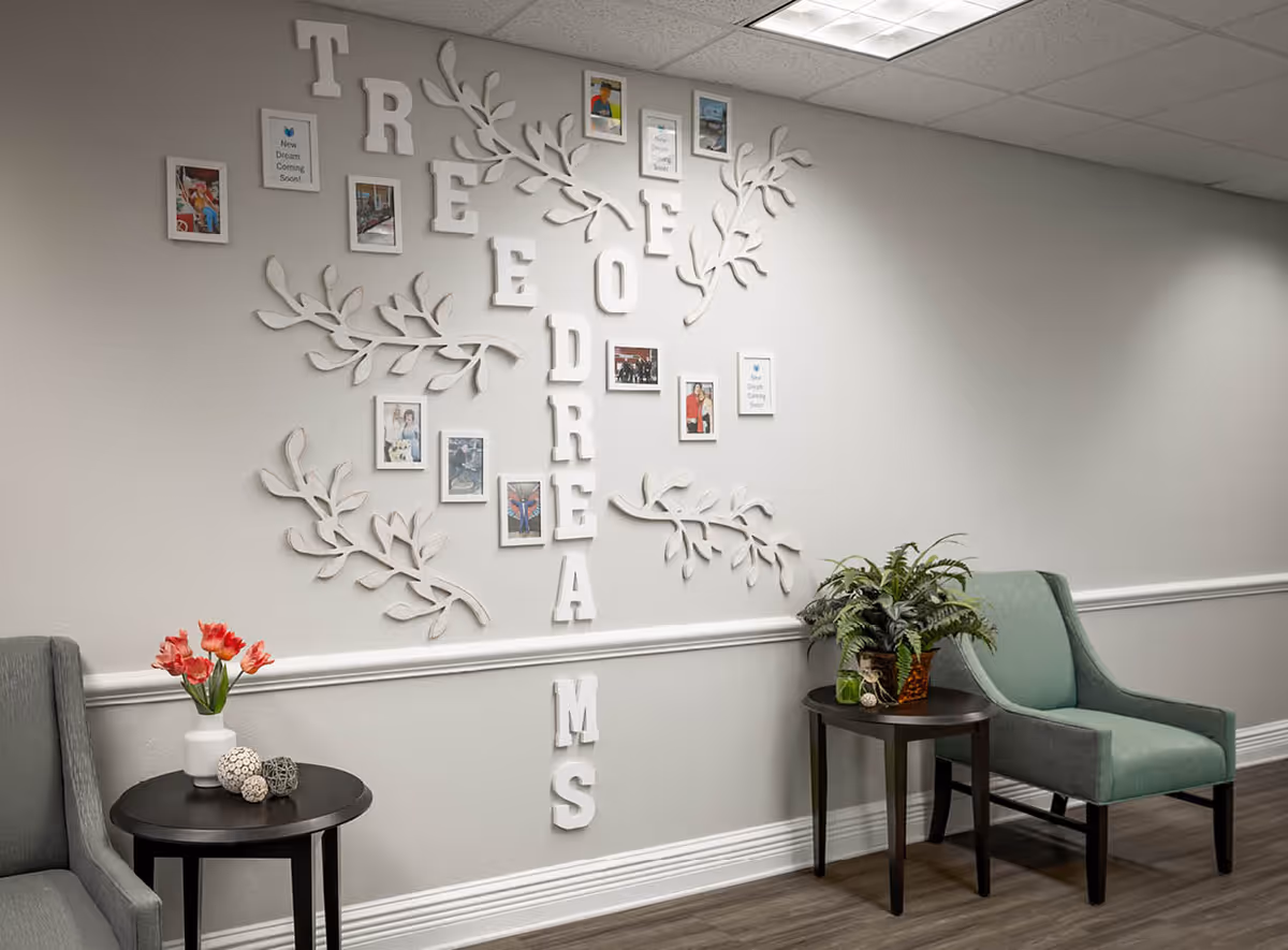 A cozy seating area with two armchairs separated by small round tables. One table has a vase with red flowers and decorative spheres, while the other has a potted plant. On the wall behind, white decorative letters and branches form the words 'TREE OF DREAMS' with framed photos and quotes interspersed.