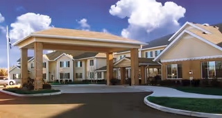 Exterior view of Winterberry Heights Senior Living community building with a large covered entrance, brick and beige siding, multiple windows, and a clear blue sky with some clouds.