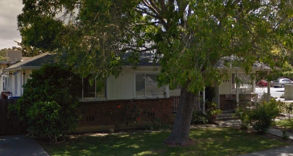 Exterior view of a single-story residential building partially obscured by a large tree and bushes, with a small front yard and a driveway on the left side.