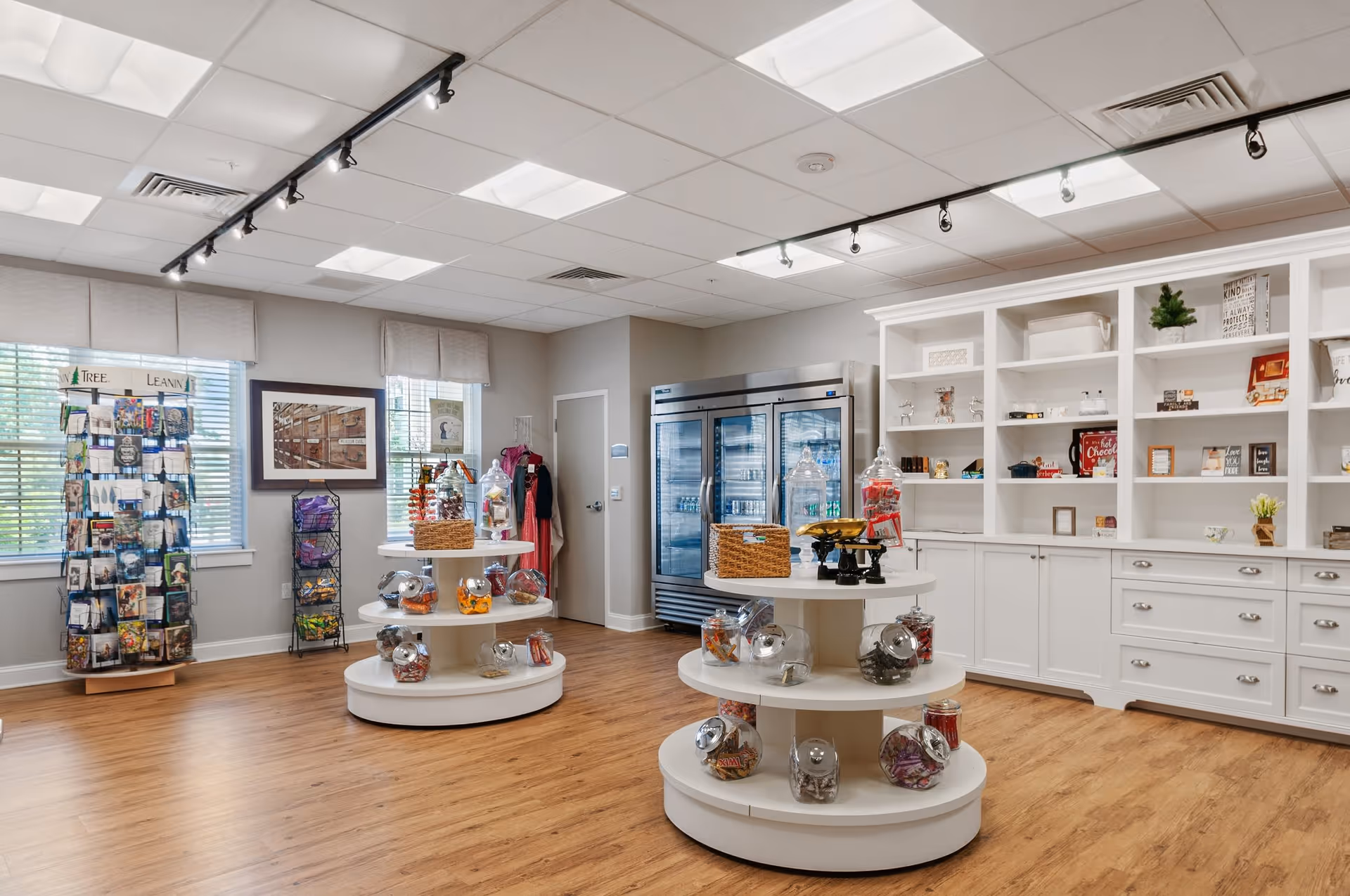 Interior view of a bright and spacious retail or gift shop area in a senior living facility. The room features wooden flooring, white shelving units filled with various decorative items and small gifts, two round display tables with glass jars containing candy or snacks, and a large refrigerated display case against the back wall. There are windows with blinds allowing natural light to enter, and racks displaying postcards or brochures near the windows.