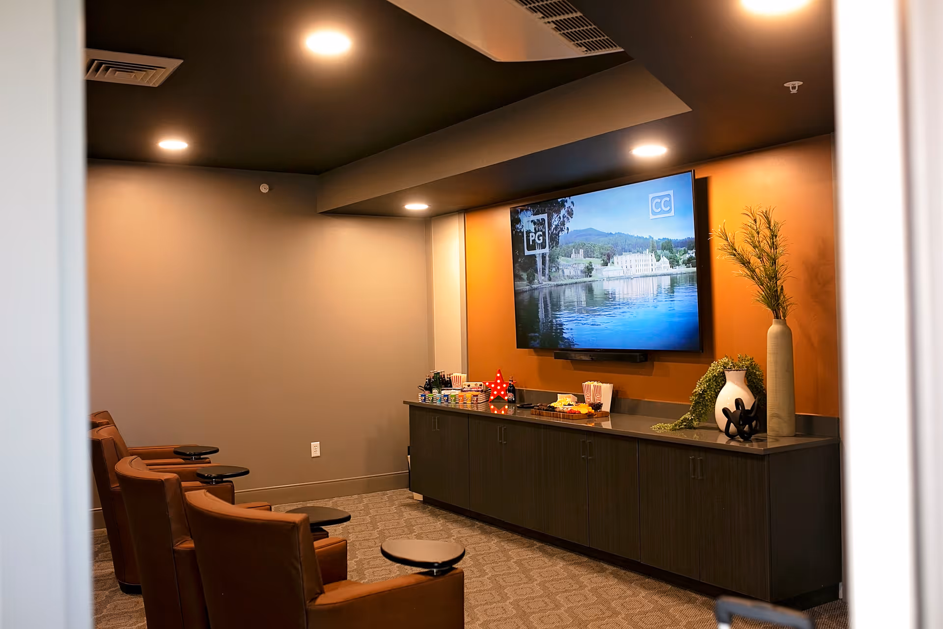A small media room with four brown leather chairs equipped with small black trays facing a large flat-screen TV mounted on an orange accent wall. Below the TV is a long dark cabinet with various snacks and decorative items, including vases with greenery. The room has recessed ceiling lights and neutral-colored walls and carpet.