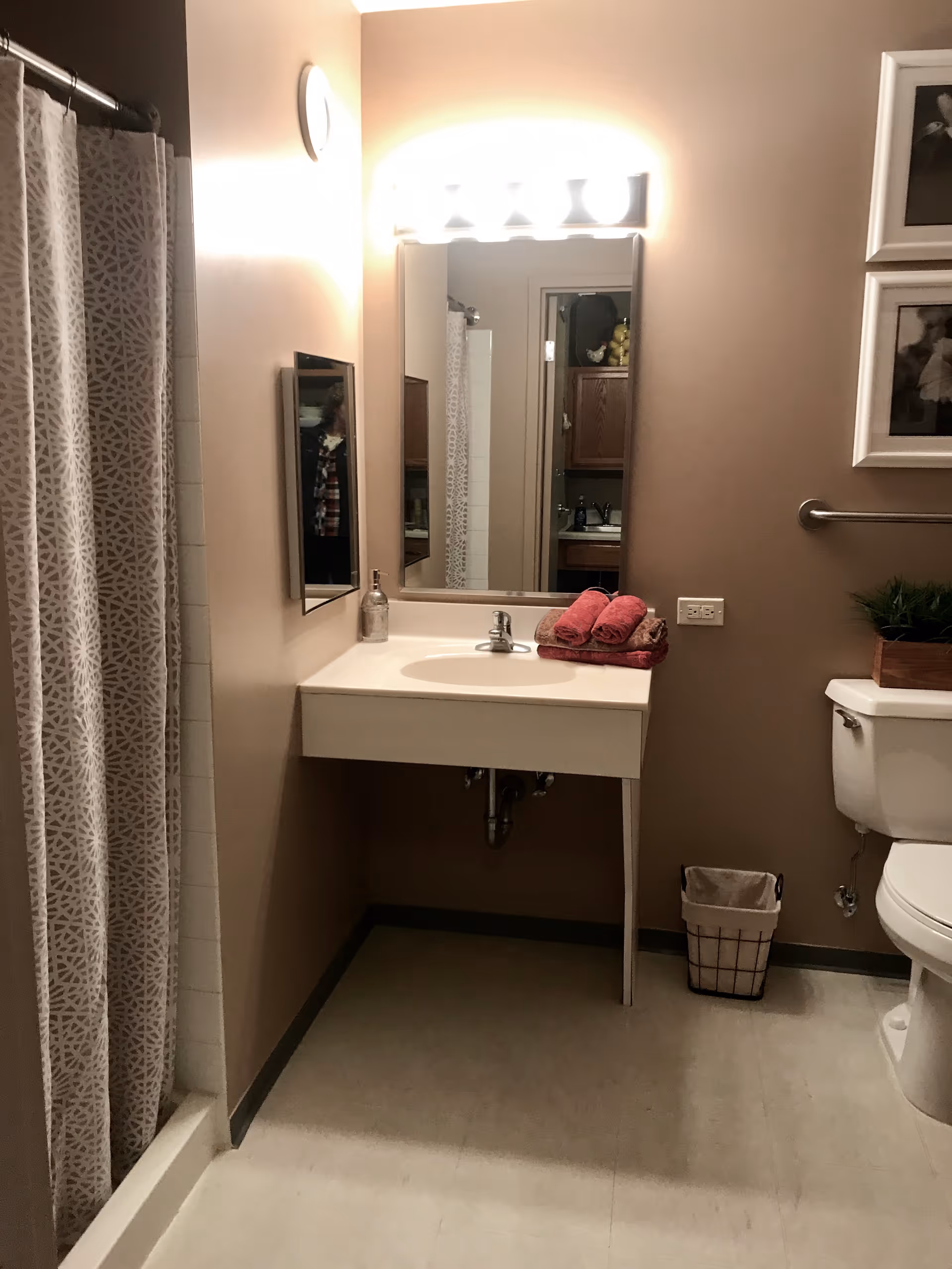 A bathroom with a white sink and countertop, a large mirror above the sink with bright lights, a shower with a patterned curtain, a toilet with a plant on top, a small trash bin on the floor, and two rolled red towels on the sink.