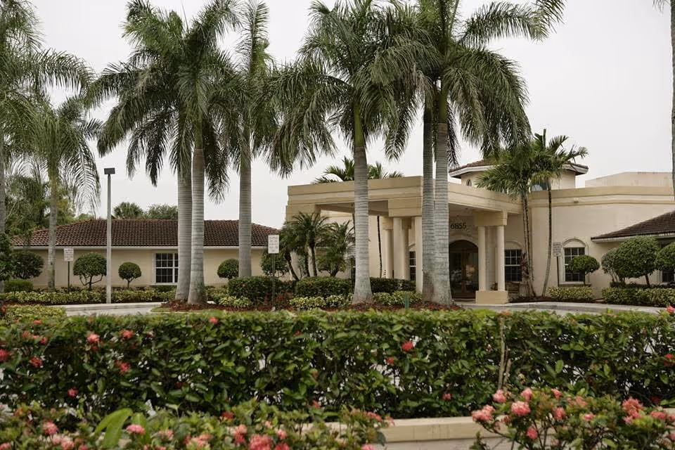 Exterior view of HarborChase of Tamarac facility showing a beige building with a covered entrance, surrounded by tall palm trees, manicured bushes, and flowering plants under an overcast sky.
