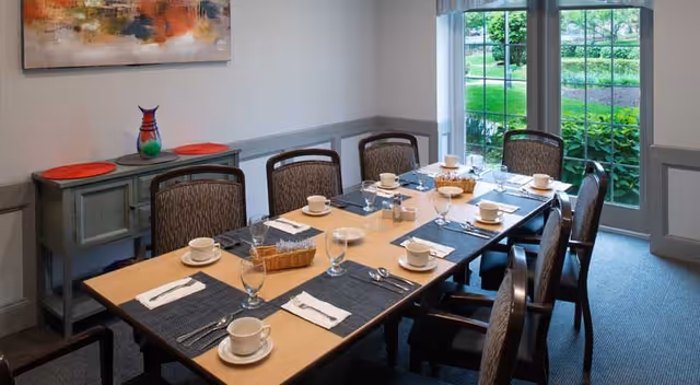 A dining room with a rectangular wooden table set for eight people. Each place setting includes a white cup and saucer, a glass, a fork, a knife, and a napkin. There are two small baskets with bread or rolls on the table. The room has a large window showing greenery outside, a sideboard with a colorful vase, and a modern abstract painting on the wall.