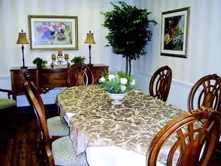 A dining room with a wooden table covered by a patterned tablecloth and surrounded by six wooden chairs with cushioned seats. There is a decorative centerpiece with white flowers on the table. In the background, there is a wooden sideboard with two lamps, framed floral artwork on the walls, and a tall potted plant in the corner.