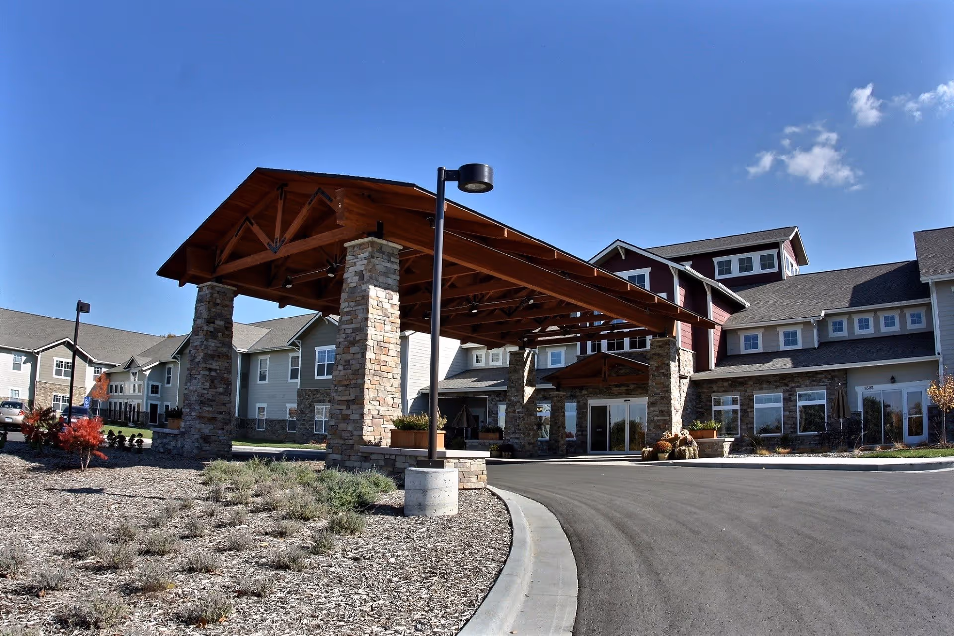 Exterior view of Westchester Village of Lenexa showing the main entrance with a large covered driveway supported by stone pillars, surrounded by landscaped grounds under a clear blue sky.