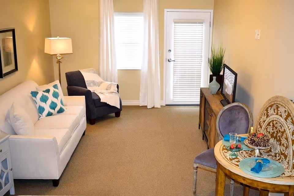 A cozy living room with a white sofa adorned with a blue and white patterned pillow, a dark armchair with a white throw blanket, a wooden sideboard with a vase and TV, and a small round dining table set with plates, glasses, and a chocolate cake. The room has beige walls, carpeted floor, a window with white curtains, and a door with blinds.