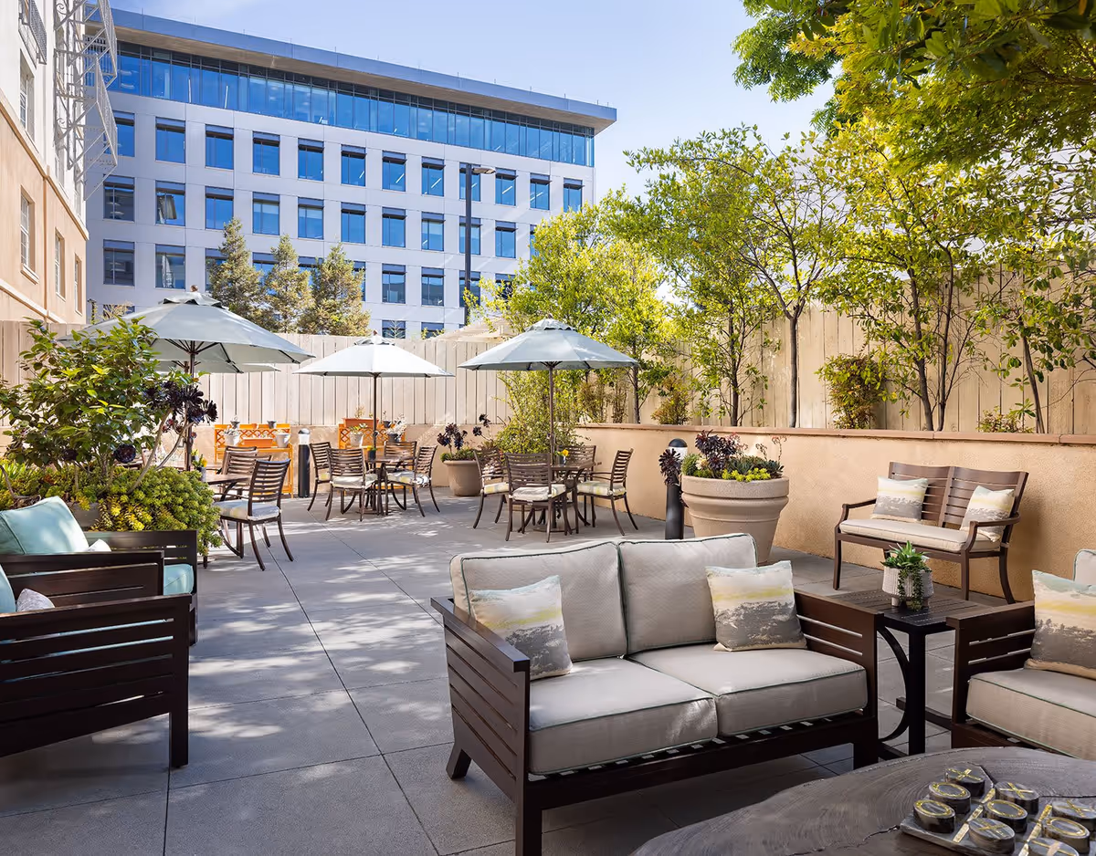 Outdoor patio area with cushioned wooden seating, tables with umbrellas, potted plants, and trees, set against a backdrop of a multi-story office building and a wooden fence.