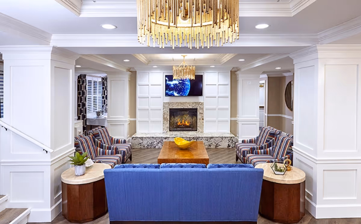 A bright and elegant living room in an assisted living facility featuring a blue sofa facing a fireplace with a mounted TV above it. Two striped armchairs flank a wooden coffee table with a yellow decorative bowl. The room has white paneled walls, recessed lighting, and a large gold chandelier hanging from the ceiling.
