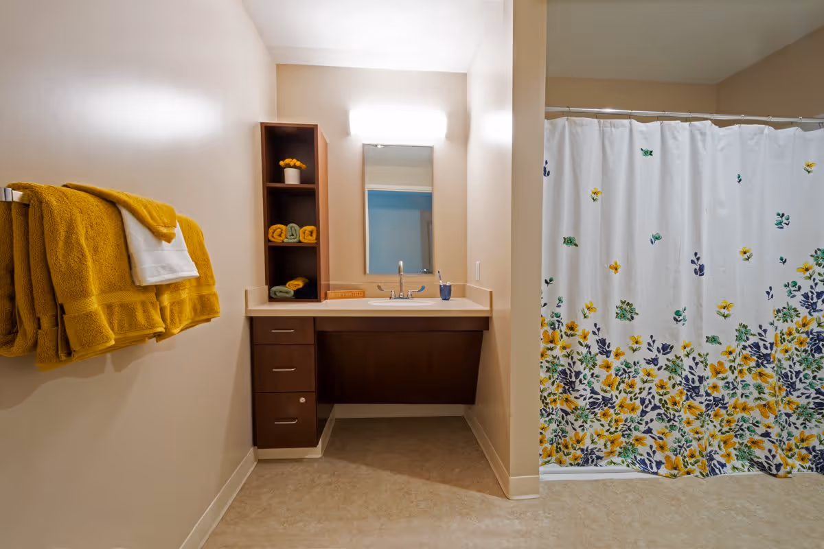 A clean bathroom with beige walls and floor. On the left wall, there are yellow and white towels hanging on a towel rack. In the center, there is a vanity with a sink, a mirror above it, and a light fixture. The vanity has dark wood cabinets and shelves with rolled towels and a small potted plant. On the right side, there is a shower area with a white curtain decorated with yellow, green, and blue floral patterns.