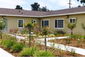 Single-story building with beige exterior walls and multiple windows, surrounded by a garden with small trees, shrubs, and a paved walkway under a partly cloudy sky.