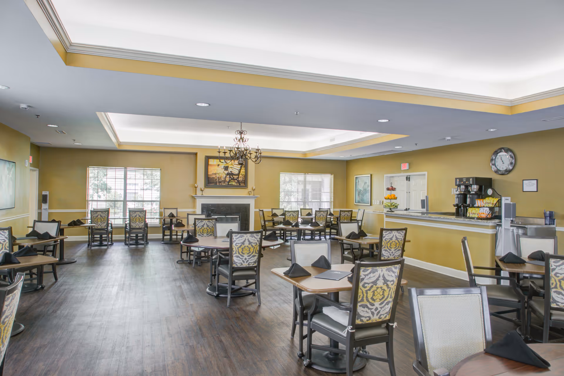 A spacious dining room with multiple round tables and chairs arranged neatly. Each table has black folded napkins and menus. The room features large windows letting in natural light, a chandelier hanging from the ceiling, a fireplace with a painting above it, and a counter area with a coffee machine and fruit display. The walls are painted in a warm yellow tone and the floor has dark wood finish.