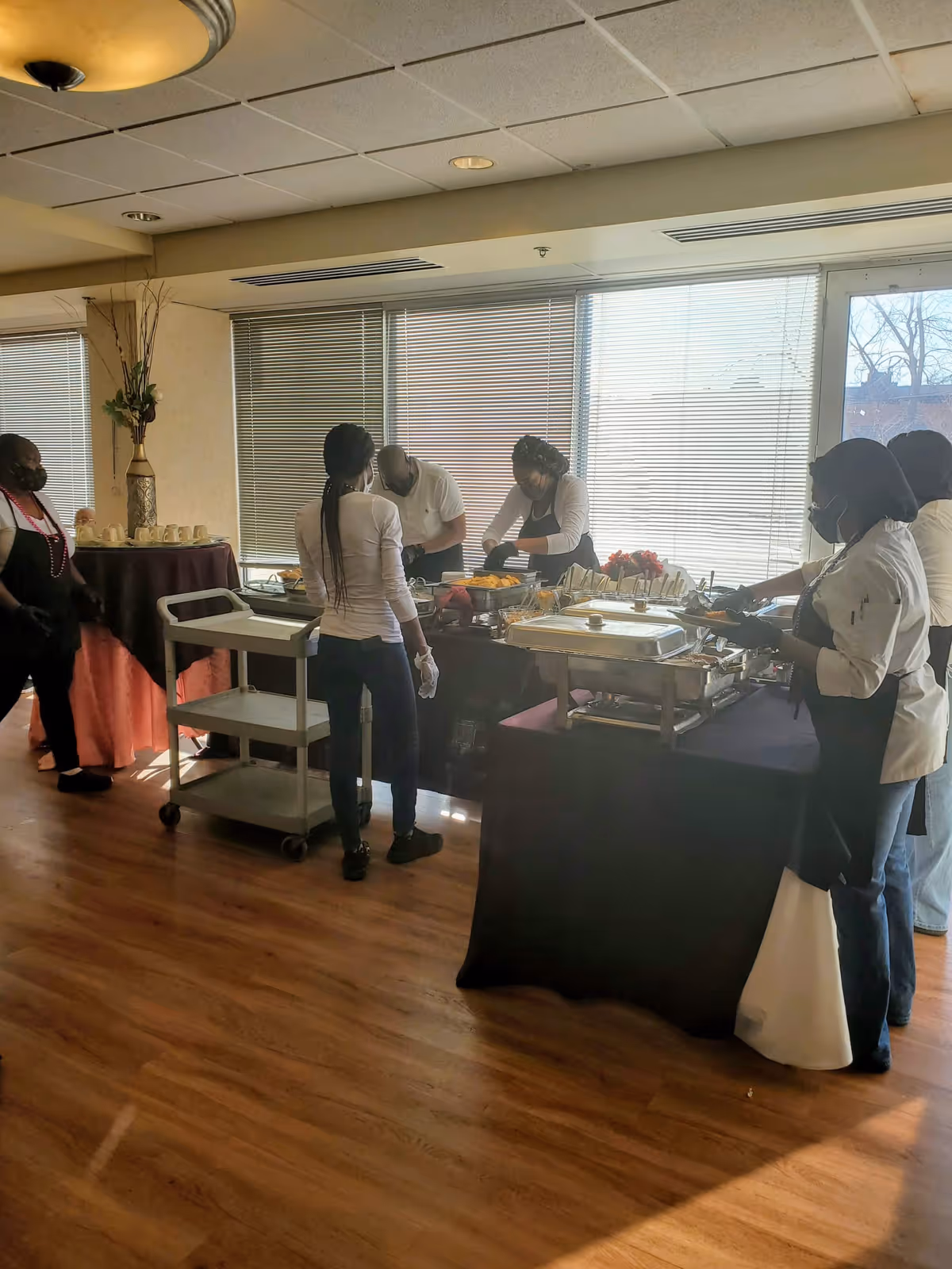 Several people wearing aprons and gloves are preparing and serving food on a buffet table in a room with large windows and wooden flooring. There is a serving cart nearby and a table with cups and a vase with flowers in the background.