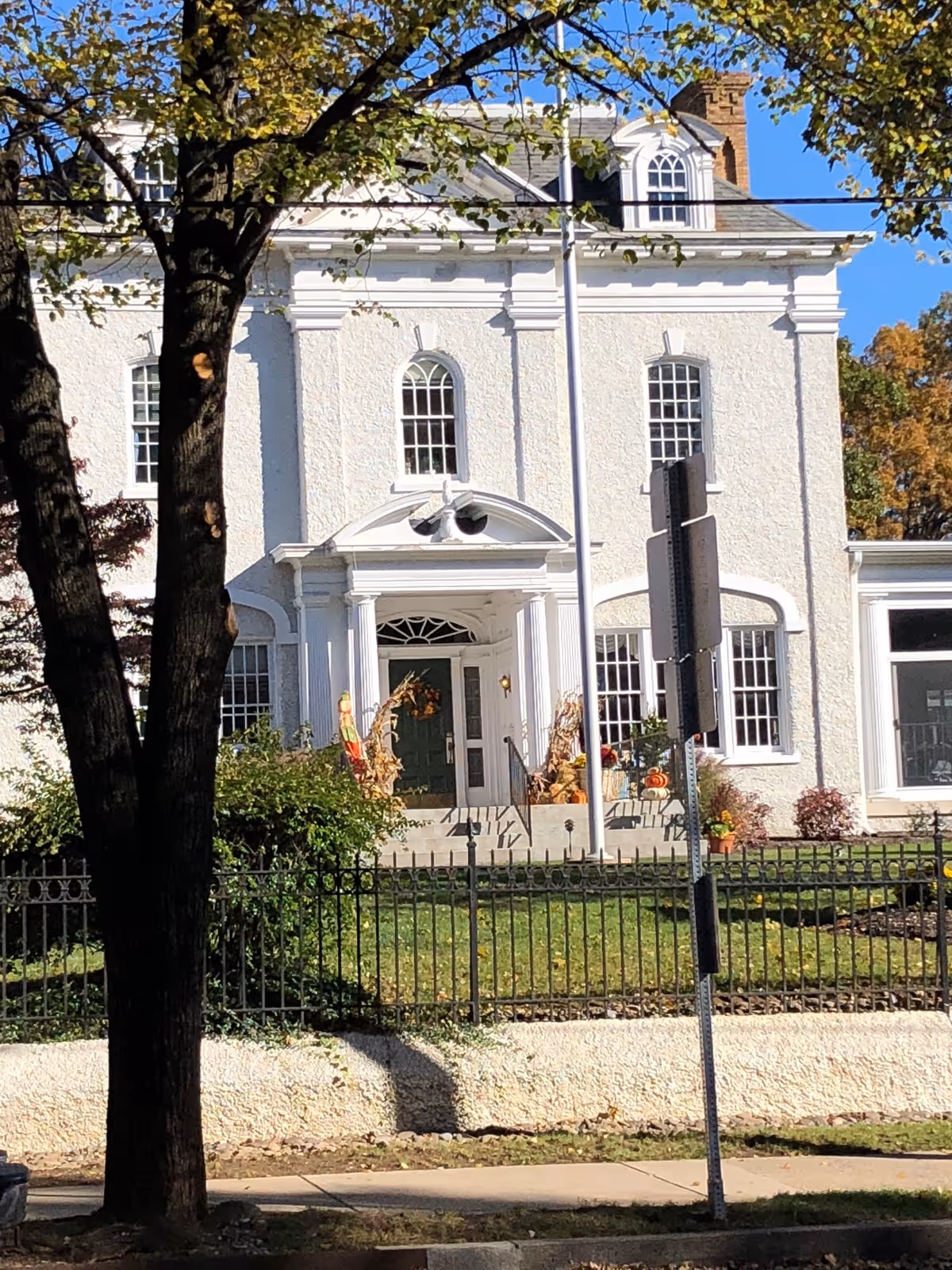 Front exterior view of a large white building with classical architectural features including columns and arched windows. There is a green door decorated with a wreath, and fall decorations such as cornstalks and pumpkins on the steps leading to the entrance. Trees and a black metal fence are visible in the foreground under a clear blue sky.