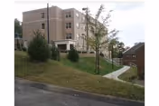 Exterior view of a multi-story beige building with several windows, situated on a grassy slope with a paved walkway leading to the entrance. There are a few trees and another smaller building visible to the right.