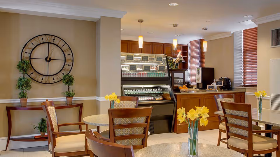 A cozy dining area in Atria Marland Place featuring round tables with wooden chairs, each table decorated with a vase of yellow flowers. In the background, there is a counter with a display case containing beverages and snacks, wooden cabinets, and windows with wooden blinds. A large decorative clock and potted plants are on the wall to the left.