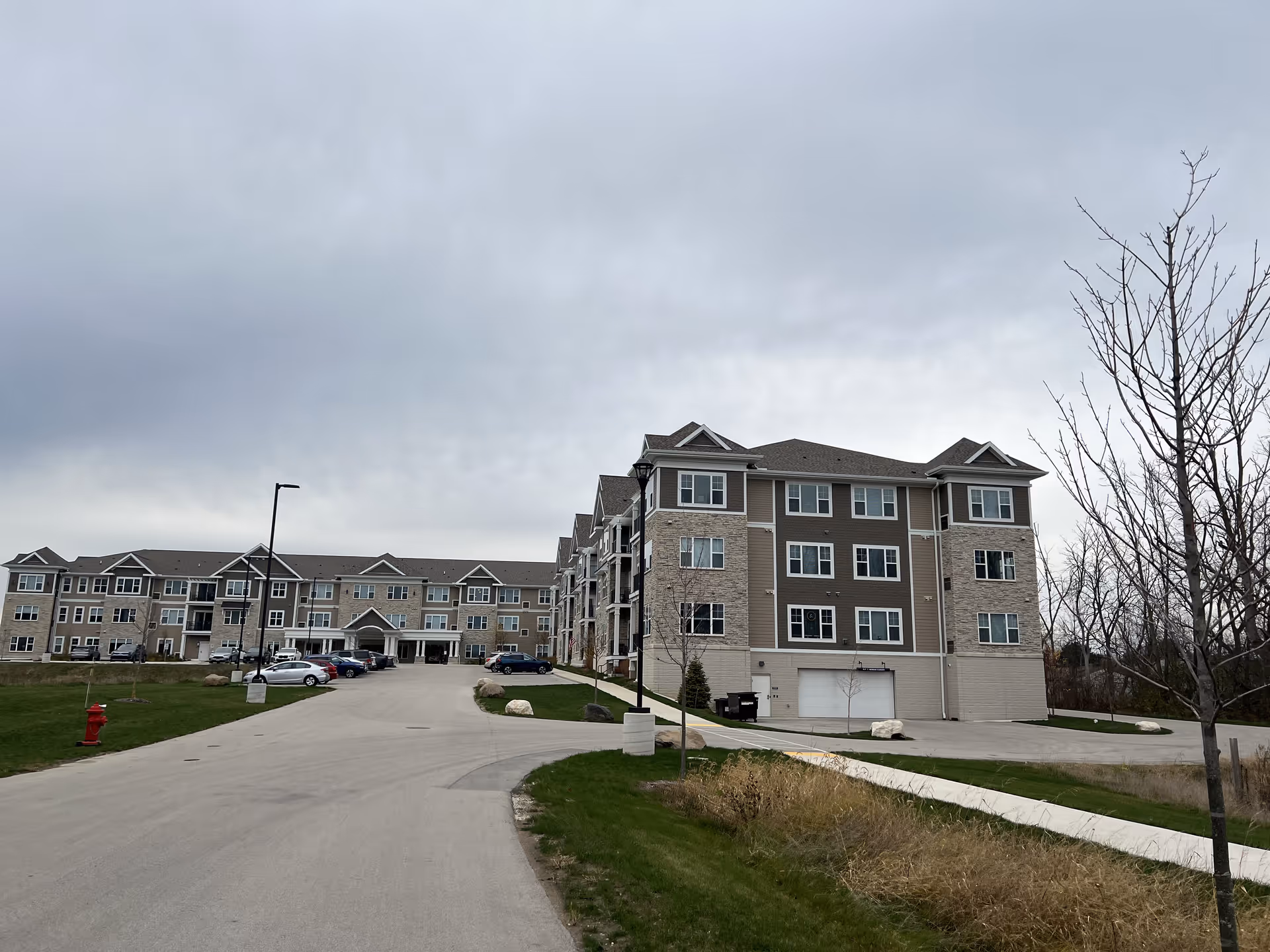 Exterior view of a large, multi-story senior living facility building with a driveway leading to the entrance. Several cars are parked near the entrance, and there are leafless trees and grassy areas surrounding the building under a cloudy sky.