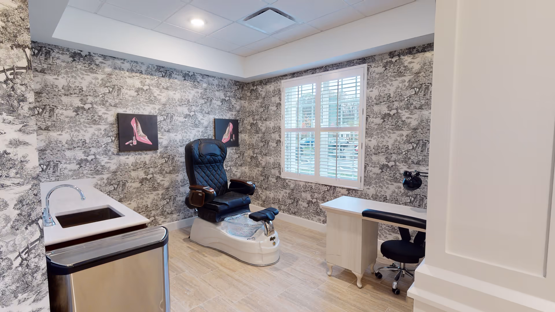 A pedicure station in a senior living facility with a black leather pedicure chair, a small desk with a black swivel chair, a sink with a faucet, and wallpaper featuring a black and white scenic pattern. Two framed pictures of pink high-heeled shoes hang on the wall, and a window with white shutters lets in natural light.