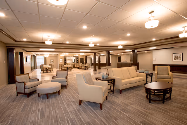 A spacious and well-lit common area in a senior living facility featuring multiple seating arrangements with beige and gray upholstered chairs, a beige sofa, round wooden coffee tables, and dining tables with chairs in the background. The room has a neutral color palette with carpeted flooring, recessed lighting, and ceiling fixtures.