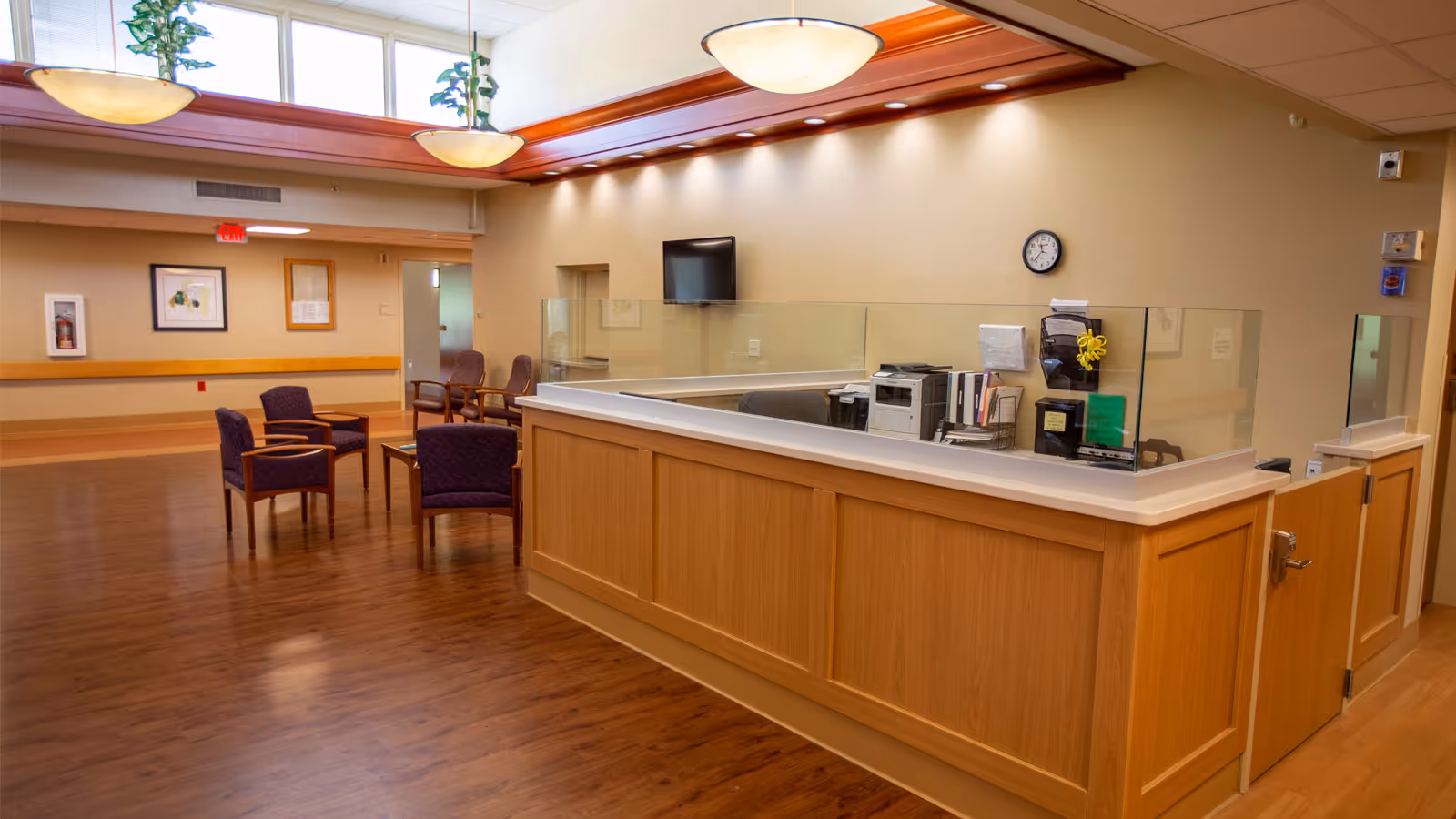 Reception area in a senior living facility with a wooden desk featuring a glass partition, office equipment, and a clock on the wall. There are several purple upholstered chairs arranged around a small table on the wooden floor. The ceiling has recessed lighting and large hanging light fixtures, with windows near the ceiling letting in natural light.