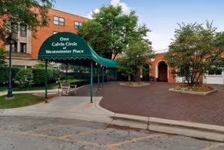 Entrance area of Westminster Place featuring a green canopy with the text 'One Calvin Circle Westminster Place', surrounded by trees and a brick-paved driveway leading to the building entrance.