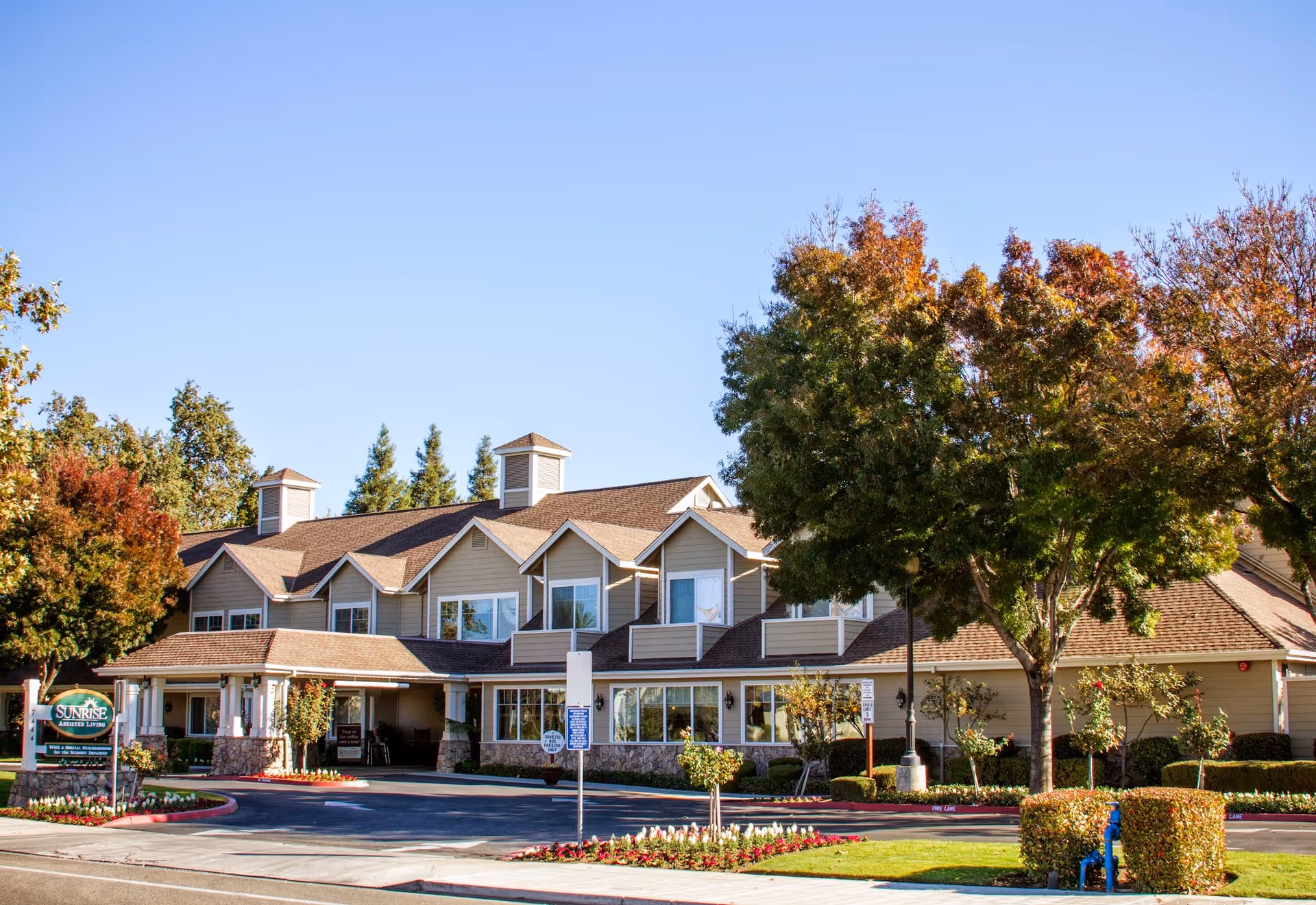 Exterior view of a senior living facility building with beige siding and brown roof, surrounded by trees with autumn foliage and landscaped flower beds under a clear blue sky.