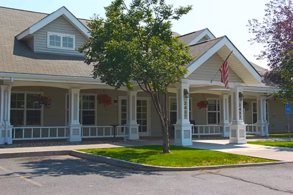 Exterior view of a single-story building with a covered porch supported by white columns, hanging flower baskets, a tree in front, and an American flag displayed near the entrance.