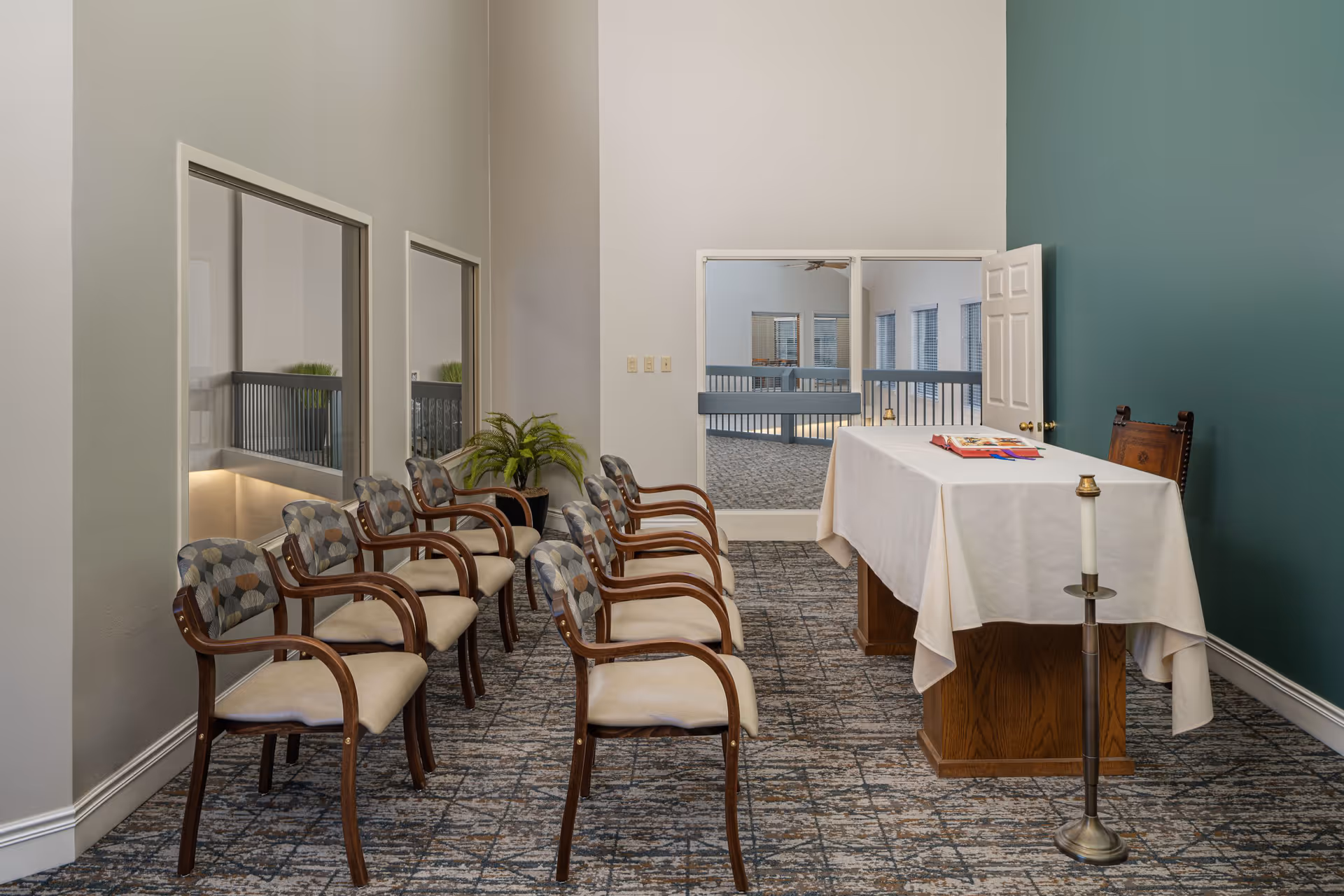 A small chapel or meditation room with a row of eight cushioned wooden chairs facing a wooden altar covered with a white cloth. On the altar, there are some books and a candle stand with a lit candle. The room has a carpeted floor, light-colored walls, and a green accent wall. There are two large windows on one side and a door with glass panels leading to an adjacent area.