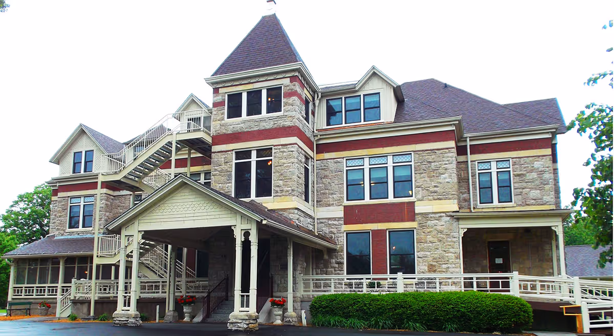 Exterior view of a large, multi-story stone building with red and beige accents, featuring multiple windows, a covered entrance with columns, an external staircase, and a ramp. The building is surrounded by greenery and trees.