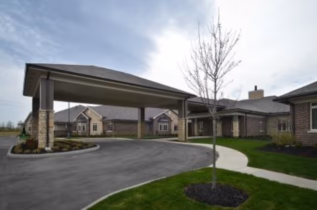 Exterior view of The Ganzhorn Suites showing a covered driveway entrance with brick pillars, a curved asphalt driveway, a small tree planted in a mulched area, and a sidewalk leading to the building entrance under a cloudy sky.