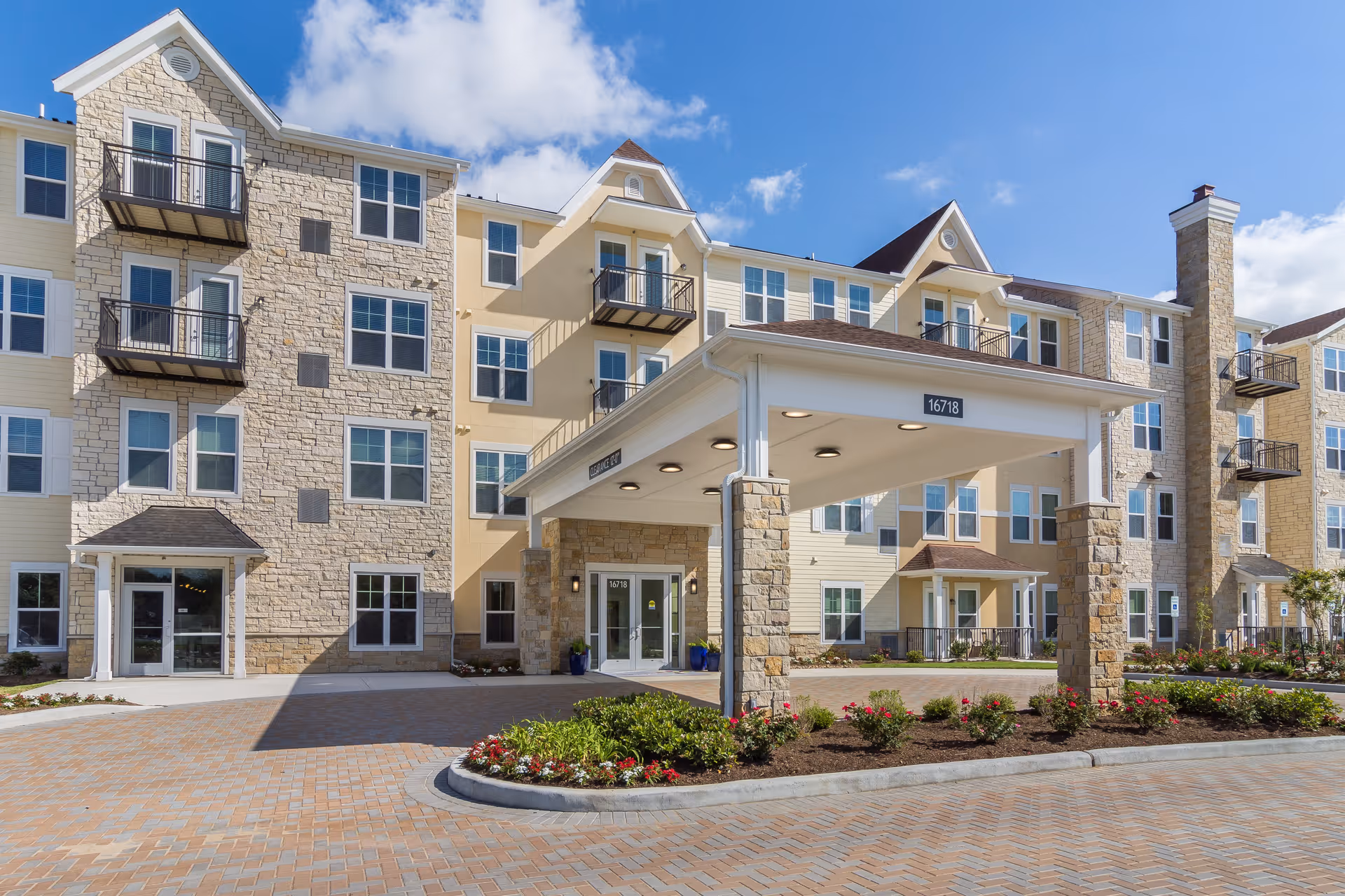 Exterior view of a multi-story senior living facility with stone and beige siding, balconies, a covered entrance with pillars, and landscaped flower beds under a blue sky with some clouds.