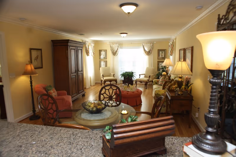 A cozy living room with wooden flooring, featuring a round glass-top table with decorative items, several upholstered chairs, a sofa with patterned cushions, a wooden armoire, multiple lamps providing warm lighting, framed artwork on the walls, and large windows with curtains allowing natural light to fill the room.
