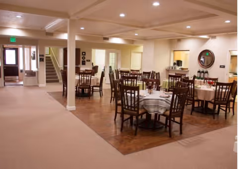 Interior view of a dining area in an assisted living facility with multiple round tables covered with tablecloths and surrounded by wooden chairs. The room has a combination of carpet and wood flooring, recessed lighting in the ceiling, and a staircase near the entrance.