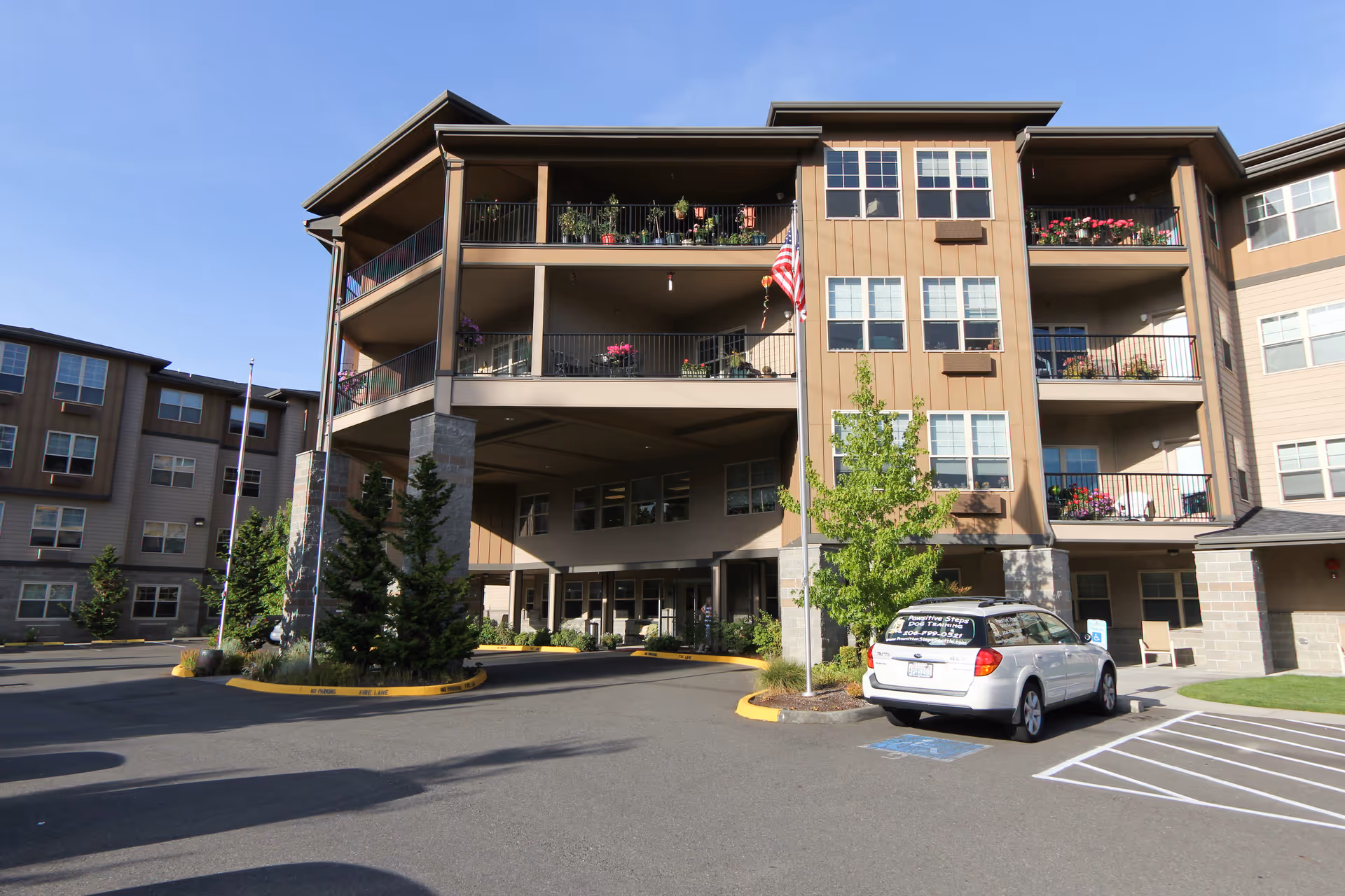 Exterior view of a multi-story senior living facility building with balconies decorated with plants and flowers. There is a driveway with a white vehicle parked in a handicapped parking space near the entrance. An American flag is displayed on a flagpole in front of the building.