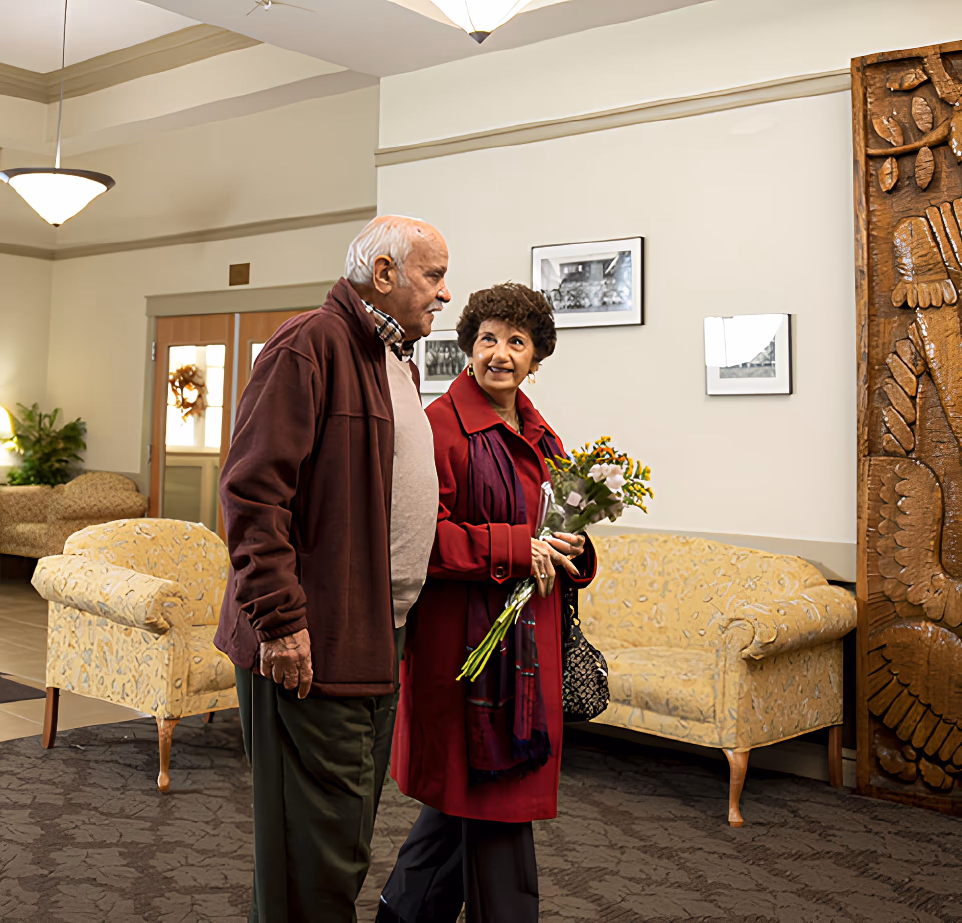 An elderly man and woman walking together inside a well-lit common area with patterned armchairs, framed black and white photos on the wall, and a carved wooden panel. The woman is holding a bouquet of flowers and looking at the man.