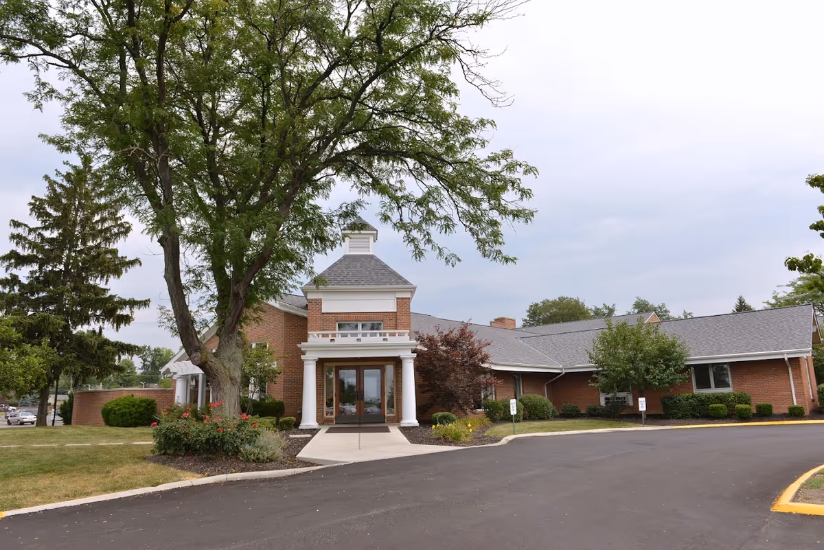 Front exterior view of a single-story brick building with a peaked roof and white columns at the entrance, surrounded by trees and landscaping with a paved driveway in front.