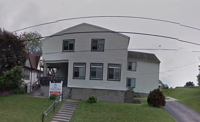 Exterior view of a two-story white building with multiple windows, a small staircase leading up to the entrance, and a grassy lawn in front. There is a sign near the stairs indicating the facility name Heartful Hands. The sky is overcast.