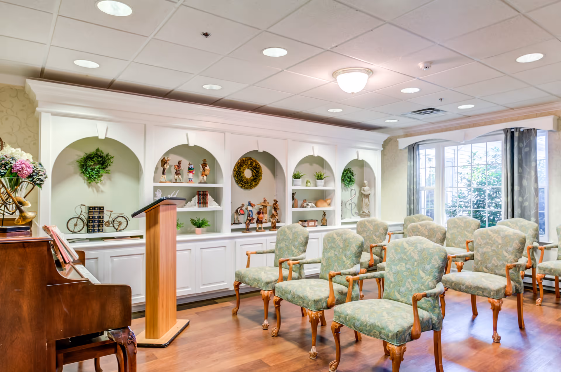 A bright room with several upholstered chairs arranged in rows facing a wooden podium and a piano. The back wall features built-in white shelves with decorative items such as figurines, plants, and wreaths. Large windows with curtains allow natural light to fill the space.