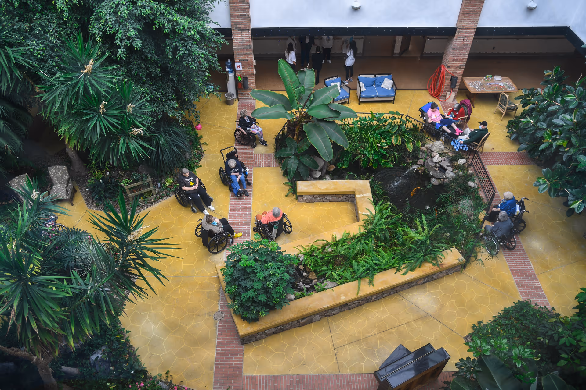 Overhead view of an indoor courtyard at a senior living facility with several elderly individuals in wheelchairs gathered around a central garden area with lush green plants and a small pond. There are seating areas with blue cushions and a few people sitting and conversing. The floor is yellow with brick accents, and the space is surrounded by tall plants and trees.